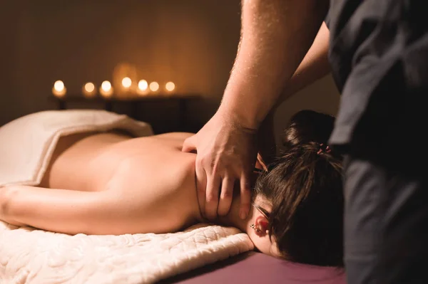 Mens hands make a therapeutic neck massage for a girl lying on a massage couch in a massage spa with dark lighting. Close-up. Dark Key — Stock Photo, Image