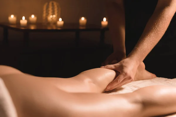 Close-up van mannelijke handen doen kalf massage van vrouwelijke benen in een donkere kamer met kaarsen op de achtergrond. Cosmetologie en wellness-behandelingen — Stockfoto