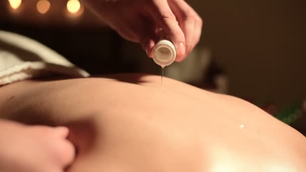 Zbliżenie: męskie dłonie w salonie uratowanych przed masażu. Olejek do masażu w ciemnym pokoju na tle płonących świec przelewa się na skórę dziewczyny — Wideo stockowe