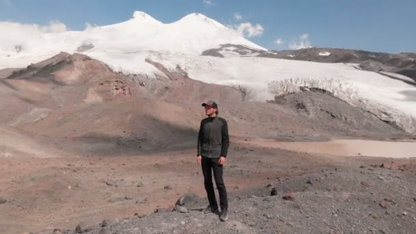 Vue ariale volant autour d'un homme debout sur une falaise dans une casquette et dans des lunettes de soleil entourées de montagnes enneigées du Caucase et de la montagne Elbrus — Video