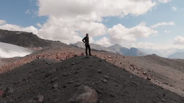 Vista arial volando alrededor de un hombre de pie sobre un acantilado en una gorra y en gafas de sol rodeado de montañas nevadas del Cáucaso y la montaña Elbrus — Vídeo de stock