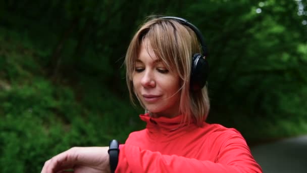 Κοντινό πλάνο του μια όμορφη ξανθιά κοπέλα στο τρέξιμο ακουστικά στέκεται στο δάσος πριν τζόκινγκ και κοιτάζοντας το ρολόι γυμναστικής. Έξυπνο ρολόι — Αρχείο Βίντεο