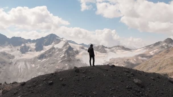 Arial Visa flyger runt en man som står på en klippa i en keps och solglasögon omgiven av snöklädda Kaukasus bergen och Elbrus berg — Stockvideo