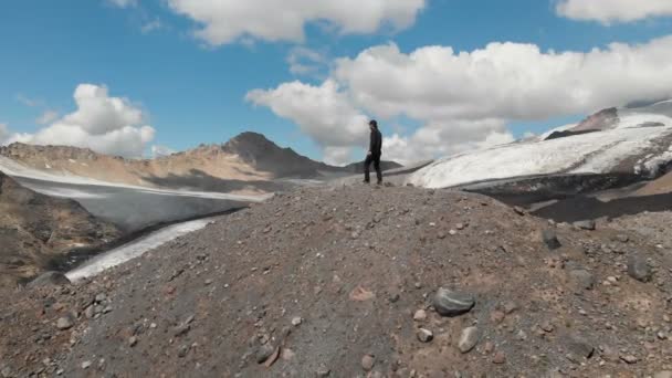 キャップとサングラス エルブラス山と雪をかぶったコーカサス山脈に囲まれた崖の上に立っている人の周りに飛んで arial ビュー — ストック動画
