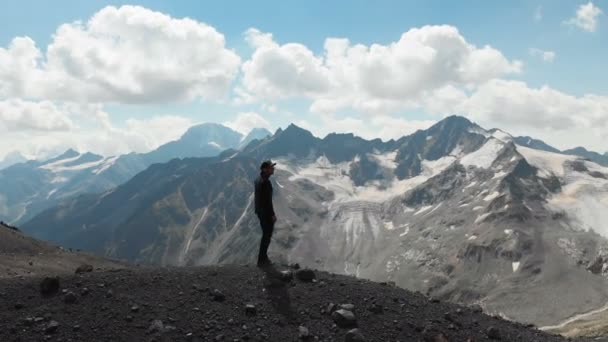 Arial widok latające wokół człowieka stojącego na klifie w czapkę i okulary przeciwsłoneczne, otoczony ośnieżonymi szczytami Kaukazu — Wideo stockowe