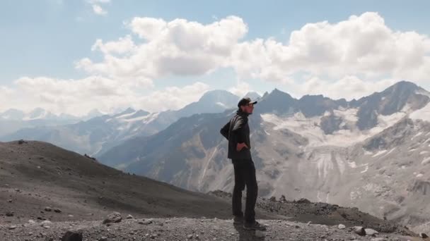 Vista arial volando alrededor de un hombre de pie sobre un acantilado en una gorra y en gafas de sol rodeado de montañas nevadas del Cáucaso — Vídeos de Stock