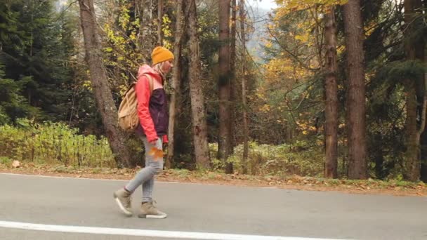 Λεπτή κοπέλα με το κίτρινο καπέλο και ένα vintage σακίδιο βόλτες κατά μήκος σε άσφαλτο στο δάσος το φθινόπωρο κίτρινου κωνοφόρων. Πλάγια όψη — Αρχείο Βίντεο