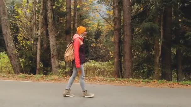 Ein junges, schlankes Mädchen mit gelbem Hut und historischem Rucksack läuft eine asphaltierte Straße im herbstgelben Nadelwald entlang. Seitenansicht — Stockvideo