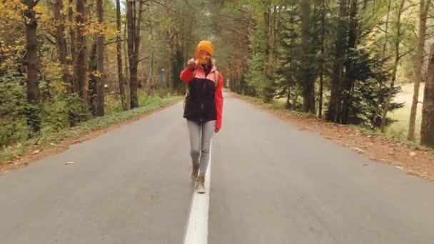Una giovane ragazza magra con un cappello giallo e uno zaino vintage cammina lungo una strada asfaltata nella foresta di conifere giallo autunno. Vista frontale — Video Stock