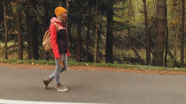 Una giovane ragazza magra con un cappello giallo e uno zaino vintage cammina lungo una strada asfaltata nella foresta di conifere giallo autunno. Vista laterale — Video Stock