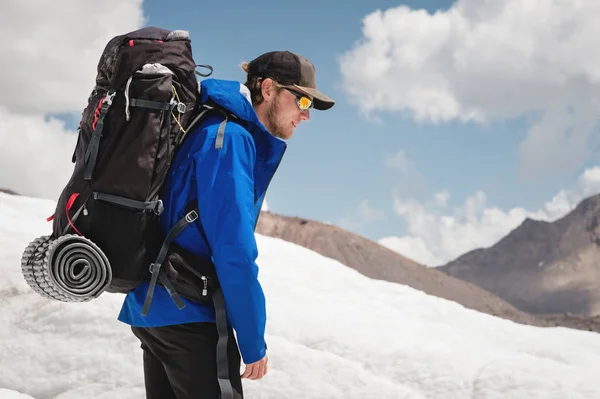 Viajante de boné e óculos de sol com uma mochila nos ombros nas montanhas nevadas no glaciar contra o céu e as nuvens. Viajante em um ambiente natural — Fotografia de Stock