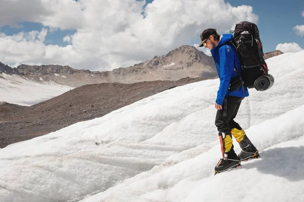 Reiziger in een pet en zonnebril met een rugzak op zijn schouders in de besneeuwde bergen op de gletsjer tegen de lucht en de wolken. Reiziger in een natuurlijke omgeving — Stockfoto