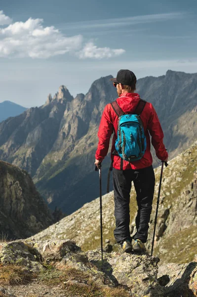 Vousatý muž v sluneční brýle a čepici s batohem stojí na vrcholu skály a dívá do vysoké skalnaté údolí v horách. Koncepce cestovního ruchu a snadný trekking v horách venkovní — Stock fotografie