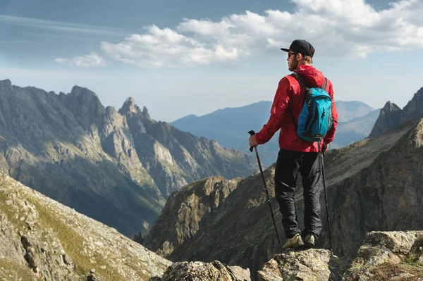 Vousatý muž v sluneční brýle a čepici s batohem stojí na vrcholu skály a dívá do vysoké skalnaté údolí v horách. Koncepce cestovního ruchu a snadný trekking v horách venkovní — Stock fotografie