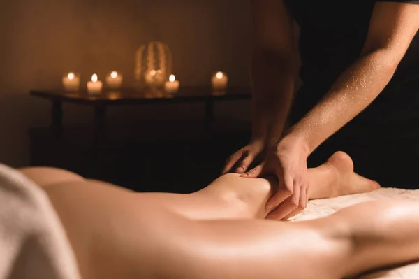 Detail z mužských rukou dělá masáž lýtek ženské nohy v temné místnosti se svíčkami na pozadí. Kosmetika a lázeňské procedury — Stock fotografie