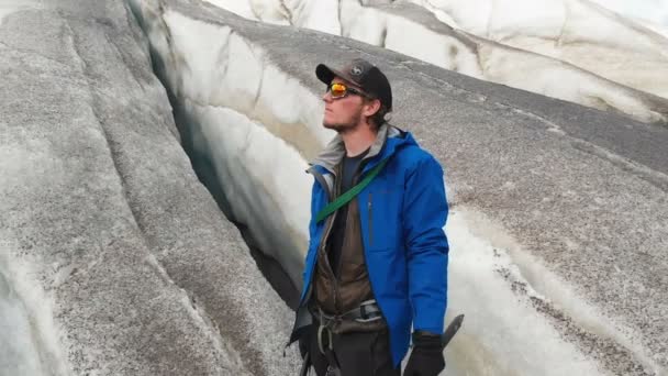 Молодой альпинист в солнцезащитных очках в кепке и колясках стоит на большом леднике с огромными трещинами в горах Кавказа. Ледник, порошок вулканической пыли — стоковое видео