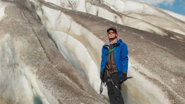 一个戴着帽子和冰爪太阳镜的年轻登山家站在一个大冰川上, 在高加索山脉上有巨大的裂缝。火山尘埃的冰川 — 图库视频影像
