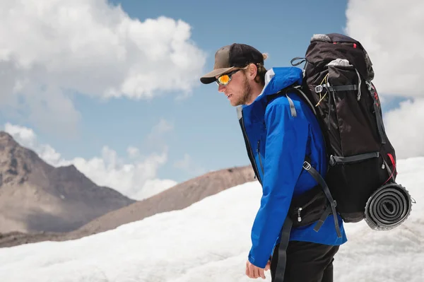 Cestovatel v čepici a brýle s batohem na ramenou v zasněžených horách na ledovci proti obloze a mraky. Cestovatel v přirozeném prostředí — Stock fotografie