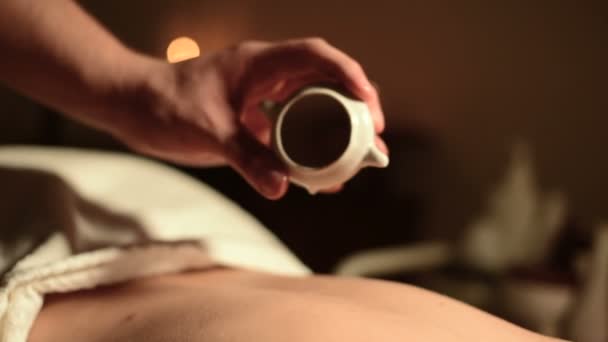 Gros plan des mains masculines dans un salon secouru avant un massage. L'huile de massage dans une pièce sombre sur le fond des bougies brûlantes est versée sur la peau des filles — Video