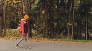 Sonbahar sarı iğne yapraklı orman içinde bir asfalt yol boyunca sarı şapka ve vintage bir sırt çantası içinde genç bir ince kız yürüyor. Yan görünüm