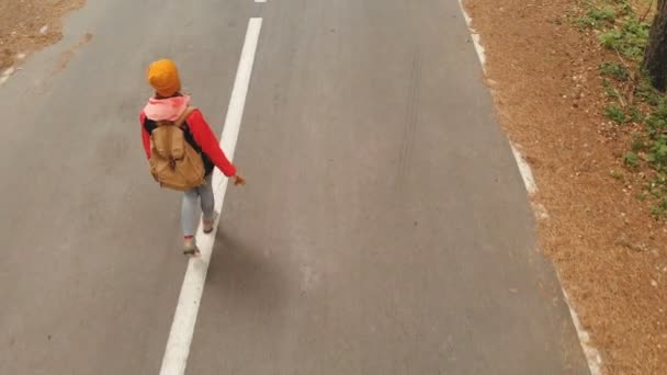 Una joven delgada con un sombrero amarillo y una mochila vintage camina a lo largo de un camino de asfalto en el bosque de coníferas amarillo de otoño. Vista trasera lateral — Vídeos de Stock