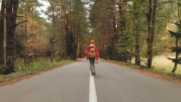 Ein junges, schlankes Mädchen mit gelbem Hut und historischem Rucksack läuft eine asphaltierte Straße im herbstgelben Nadelwald entlang. zurück — Stockvideo