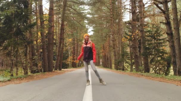 Ein junges, schlankes Mädchen mit gelbem Hut und historischem Rucksack läuft eine asphaltierte Straße im herbstgelben Nadelwald entlang. Frontansicht — Stockvideo
