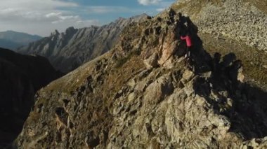 Bir sakallı bir dağcı bir şapka ve güneş gözlüğü, Genç bir adam dağlarda yüksek kayalık bir sırt tırmanıyor. Parkour dağlarda. Arial görünümü
