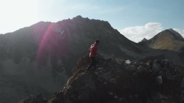 Bir sakallı bir dağcı bir şapka ve güneş gözlüğü, Genç bir adam dağlarda yüksek kayalık bir sırt tırmanıyor. Parkour dağlarda. Arial görünümü.