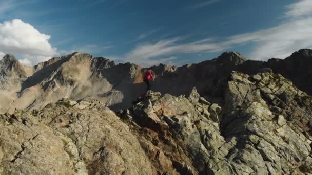 En ung kille med skägg, en bergsbestigare i en keps och solglasögon, klättrar en stenig ås högt i bergen. Parkour i bergen. Arial Visa — Stockvideo