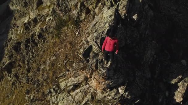 Un giovane hipster con la barba, uno scalatore con berretto e occhiali da sole, è in piedi su una cresta rocciosa alta in montagna e si guarda intorno. Parkour in montagna. Vista aerea. Movimenti dal — Video Stock