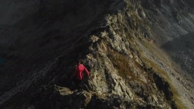 Bir sakallı bir dağcı bir şapka ve güneş gözlüğü, Genç bir adam dağlarda yüksek kayalık bir sırt tırmanıyor. Parkour dağlarda. Arial görünümü