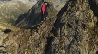 Bir sakallı bir dağcı bir şapka ve güneş gözlüğü, Genç bir adam dağlarda yüksek kayalık bir sırt tırmanıyor. Parkour dağlarda. Arial görünümü
