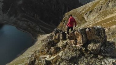 Bir sakallı bir dağcı bir şapka ve güneş gözlüğü, Genç bir adam dağlarda yüksek kayalık bir sırt tırmanıyor. Parkour dağlarda. Arial görünümü