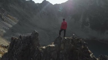 Bir sakallı bir genç hipster adam bir tırmanıcı bir şapka ve güneş gözlüğü dağlarda yüksek kayalık bir sırt üzerinde duran ve etrafa. Parkour dağlarda. Airview görünümü. Hareketi çevresinde
