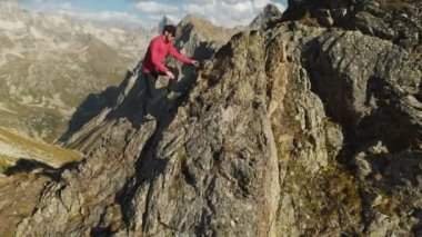 Bir sakallı bir dağcı bir şapka ve güneş gözlüğü, Genç bir adam dağlarda yüksek kayalık bir sırt tırmanıyor. Parkour dağlarda. Arial görünümü