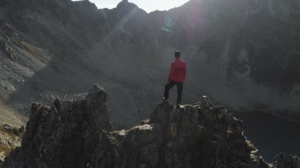 Молодой хипстер с бородой альпинист в кепке и солнцезащитных очках стоит на скалистом хребте высоко в горах и смотрит вокруг. Паркур в горах. Вид с воздуха. Движение вокруг — стоковое видео