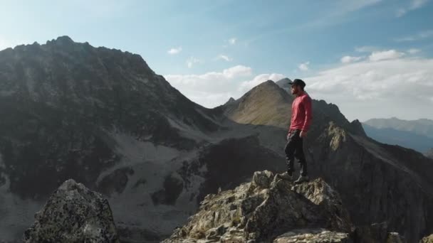 Ein junger Hipster mit Bart, Kappe und Sonnenbrille steht auf einem Felsgrat hoch in den Bergen und schaut sich um. Parkour in den Bergen. Ansicht aus der Luft. Bewegung um sich — Stockvideo