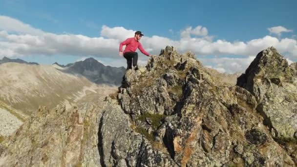 Un joven con barba, un montañero con gorra y gafas de sol, sube a una cresta rocosa en lo alto de las montañas. Parkour en las montañas. Opinión Arial — Vídeo de stock