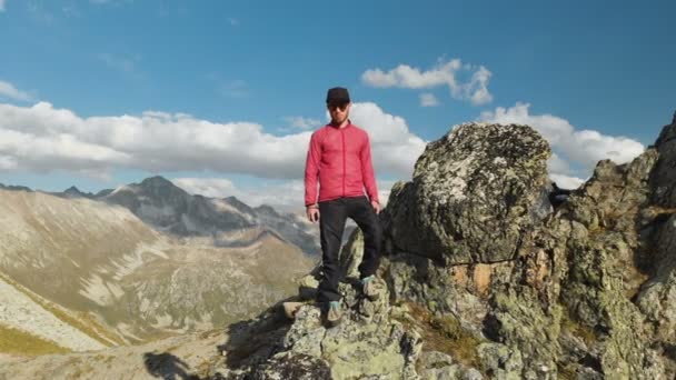 Ένας τύπος νεαρός hipster με μια γενειάδα ορειβάτης σε ένα καπέλο και γυαλιά ηλίου είναι στέκεται πάνω σε ένα βραχώδη κορυφογραμμή και η υψηλή στα βουνά και κοιτάζοντας γύρω. Parkour στα βουνά. Airview άποψη. Κίνηση από την — Αρχείο Βίντεο