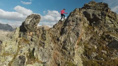 Bir sakallı bir dağcı bir şapka ve güneş gözlüğü, Genç bir adam dağlarda yüksek kayalık bir sırt tırmanıyor. Parkour dağlarda. Arial görünümü