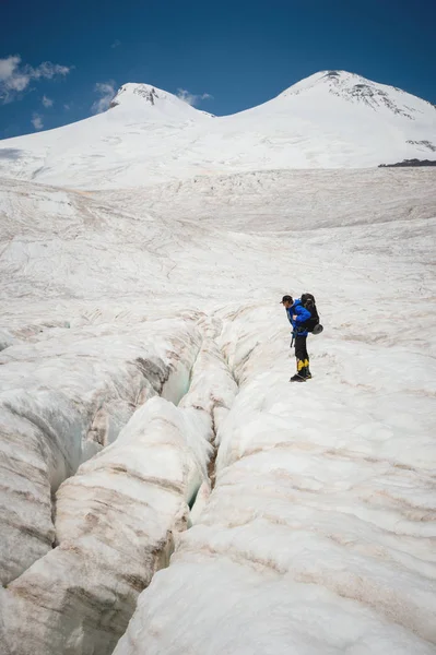 Viajero con gorra y gafas de sol con una mochila sobre sus hombros en las montañas nevadas del glaciar contra el cielo y las nubes. Viajero en un entorno natural —  Fotos de Stock