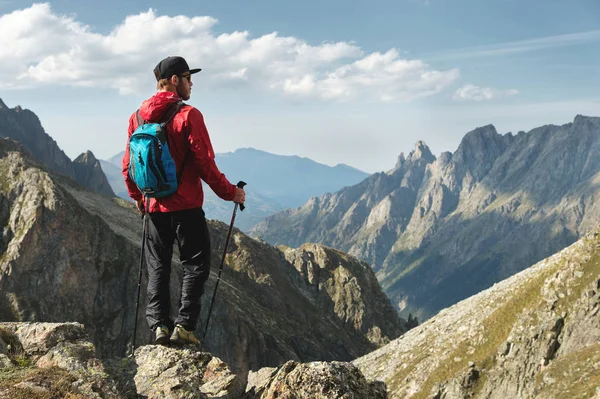 Vousatý muž v sluneční brýle a čepici s batohem stojí na vrcholu skály a dívá do vysoké skalnaté údolí v horách. Koncepce cestovního ruchu a snadný trekking v horách venkovní — Stock fotografie