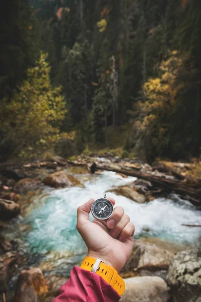 Uma bela mão masculina com uma pulseira de relógio amarela segura uma bússola magnética em uma floresta de outono conífera contra um rio de montanha com pedras rochosas. O conceito de encontrar o caminho e a verdade — Fotografia de Stock