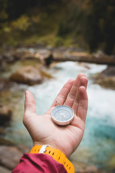 Uma bela mão masculina com uma pulseira de relógio amarela segura uma bússola magnética em uma floresta de outono conífera contra um rio de montanha com pedras rochosas. O conceito de encontrar o caminho e a verdade — Fotografia de Stock