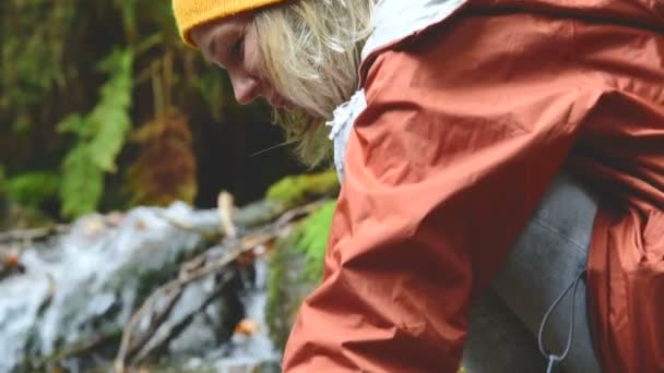 Hezká dívka cestovatel v čepici sedí na břehu horské bystřiny v jehličnatých lesů a pití čisté vody. Detailní záběr záběr — Stock video
