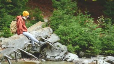 Güzel kız turistik bir şapka ve iğne yapraklı orman içinde bir dağ nehir boyunca yalan bir günlük oturan bir sırt çantası ile. Caucasus rezerv Turizm