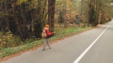 Sonbahar sarı iğne yapraklı orman içinde bir asfalt yol boyunca sarı şapka ve vintage bir sırt çantası içinde genç bir ince kız yürüyor. Yan geri görüntülemek.