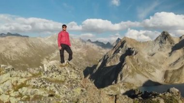 Bir sakallı bir genç hipster adam bir tırmanıcı bir şapka ve güneş gözlüğü dağlarda yüksek kayalık bir sırt üzerinde duran ve etrafa. Parkour dağlarda. Airview görünümü. Hareketi çevresinde