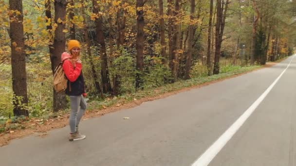 Młoda, szczupła dziewczyna w żółtym kapeluszu i plecak vintage spacery wzdłuż drogi asfaltowej w jesień żółty lasu iglastego. Widok z boku powrót — Wideo stockowe